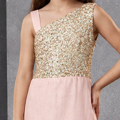 A girl wearing a one-shoulder formal dress with a shimmering gold sequined bodice and a soft blush pink skirt, shown against a textured gray background.