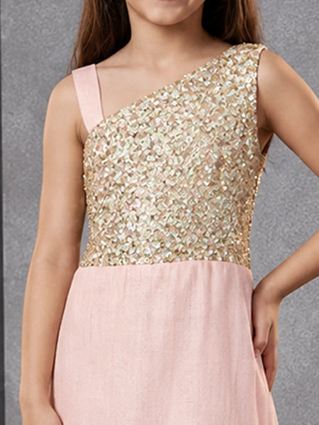 A girl wearing a one-shoulder formal dress with a shimmering gold sequined bodice and a soft blush pink skirt, shown against a textured gray background.