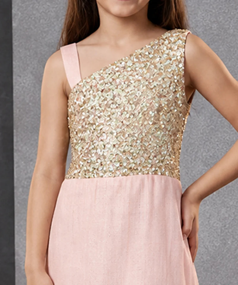 A girl wearing a one-shoulder formal dress with a shimmering gold sequined bodice and a soft blush pink skirt, shown against a textured gray background.