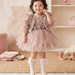 A toddler viewed from behind wearing a dusty pink ruffled tulle dress with puffed sleeves and a bow at the waist, paired with white lace socks and white shoes, standing in a soft pink nursery setting.
