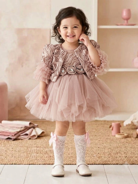 A toddler viewed from behind wearing a dusty pink ruffled tulle dress with puffed sleeves and a bow at the waist, paired with white lace socks and white shoes, standing in a soft pink nursery setting.