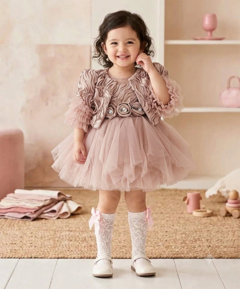 A toddler viewed from behind wearing a dusty pink ruffled tulle dress with puffed sleeves and a bow at the waist, paired with white lace socks and white shoes, standing in a soft pink nursery setting.