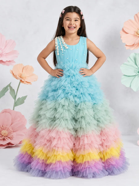 A young girl wearing a voluminous tiered tulle dress with pastel rainbow stripes, standing against a white studio backdrop decorated with large paper flowers.