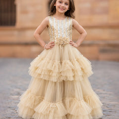 A young girl stands outdoors wearing a sleeveless champagne-gold ball gown with a heavily embellished beaded bodice and a full tiered tulle skirt. The background shows a blurred stone building and cobblestone ground.