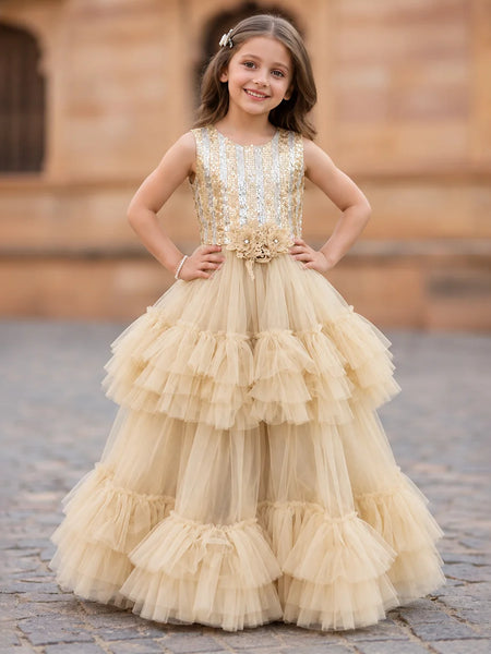 A young girl stands outdoors wearing a sleeveless champagne-gold ball gown with a heavily embellished beaded bodice and a full tiered tulle skirt. The background shows a blurred stone building and cobblestone ground.