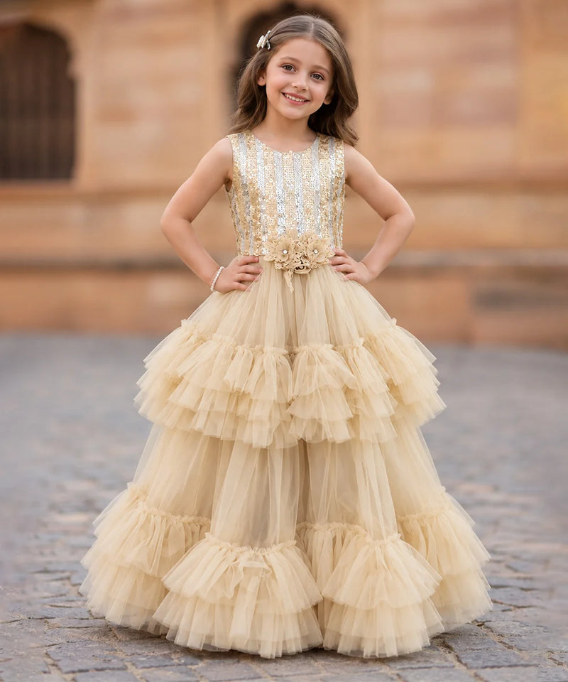 A young girl stands outdoors wearing a sleeveless champagne-gold ball gown with a heavily embellished beaded bodice and a full tiered tulle skirt. The background shows a blurred stone building and cobblestone ground.