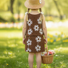 Young girl in a brown dress with white floral patterns, holding a basket of flowers, standing in a grassy field.