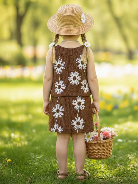 Young girl in a brown dress with white floral patterns, holding a basket of flowers, standing in a grassy field.