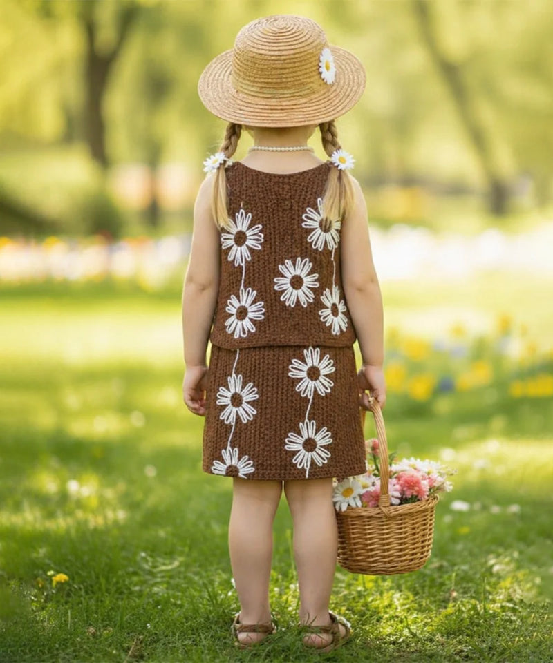 Young girl in a brown dress with white floral patterns, holding a basket of flowers, standing in a grassy field.