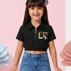 A young girl with dark hair wearing a black short-sleeve crop top with a collar, a front zipper, and a graphic '84' on the chest, paired with blue jeans, posing against a pink background with decorative flower elements.