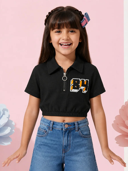 A young girl with dark hair wearing a black short-sleeve crop top with a collar, a front zipper, and a graphic '84' on the chest, paired with blue jeans, posing against a pink background with decorative flower elements.