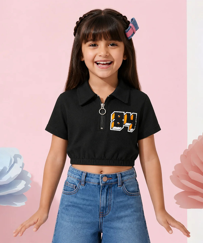 A young girl with dark hair wearing a black short-sleeve crop top with a collar, a front zipper, and a graphic '84' on the chest, paired with blue jeans, posing against a pink background with decorative flower elements.