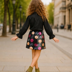 A young girl with long curly brown hair is seen from behind, wearing a black long-sleeve blouse and a black mini skirt with colorful floral patterns, paired with olive green ankle boots, walking on a city street.