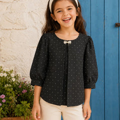 A young girl smiling and wearing a black long-sleeve top with small white polka dots, featuring a bow detail at the neckline, paired with cream pants and a cream headband, standing in front of a blue wooden door and a white stucco wall.