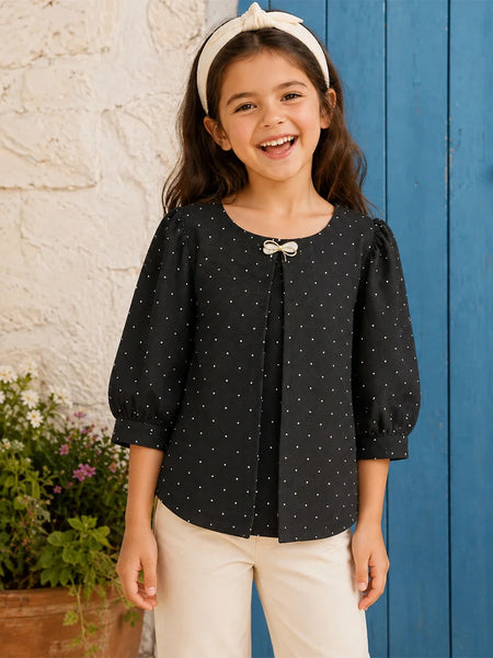 A young girl smiling and wearing a black long-sleeve top with small white polka dots, featuring a bow detail at the neckline, paired with cream pants and a cream headband, standing in front of a blue wooden door and a white stucco wall.