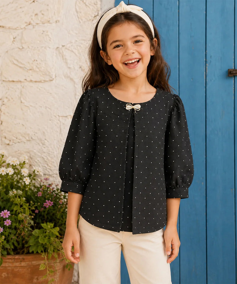 A young girl smiling and wearing a black long-sleeve top with small white polka dots, featuring a bow detail at the neckline, paired with cream pants and a cream headband, standing in front of a blue wooden door and a white stucco wall.