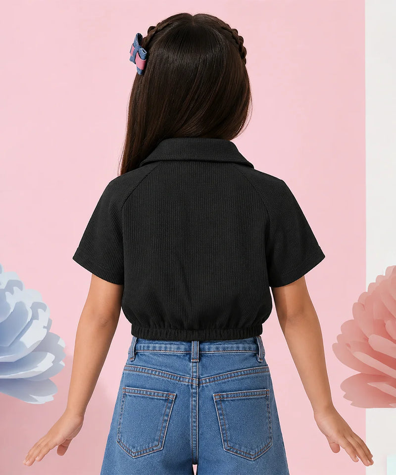 A child model shown from the back wearing a black short-sleeve collared shirt and blue jeans, standing against a pink background with decorative paper flowers.