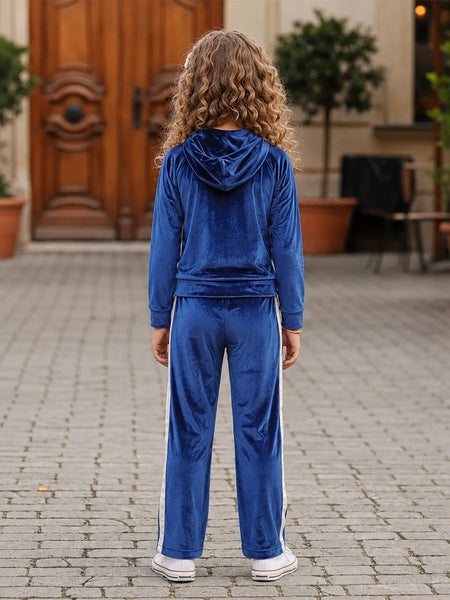 A child viewed from behind wearing a royal blue velour tracksuit with white side stripes, standing on a paved courtyard in front of a building with a wooden door.