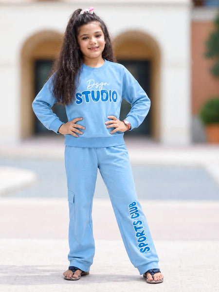 A young girl with long dark hair stands outdoors wearing a light blue matching sweatshirt and jogger pants set. The sweatshirt has 'Doge STUDIO' printed across the chest, and the jogger pants have 'SPORTS JUMPMAN' printed down the leg.