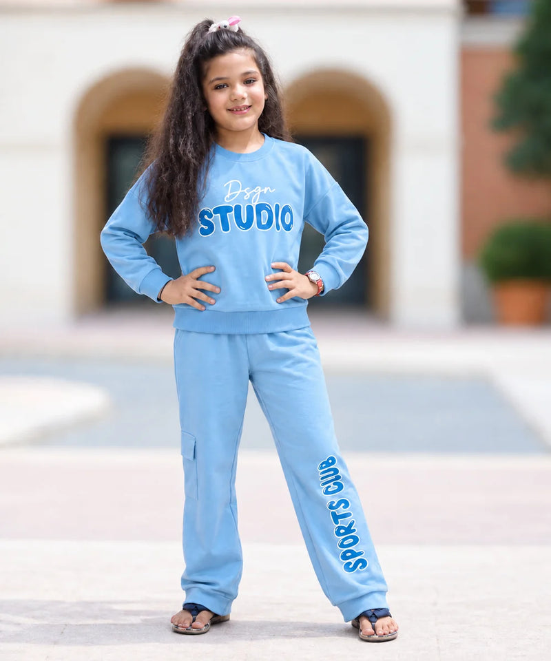 A young girl with long dark hair stands outdoors wearing a light blue matching sweatshirt and jogger pants set. The sweatshirt has 'Doge STUDIO' printed across the chest, and the jogger pants have 'SPORTS JUMPMAN' printed down the leg.