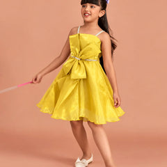 A young girl with dark hair wearing a bright yellow sleeveless dress with a large bow at the waist, white shoes, and a purple hair bow, standing against a solid peach-colored background.