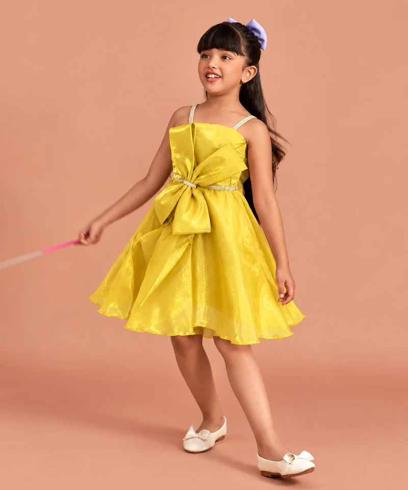 A young girl with dark hair wearing a bright yellow sleeveless dress with a large bow at the waist, white shoes, and a purple hair bow, standing against a solid peach-colored background.