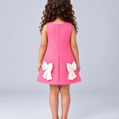 A young girl viewed from behind wearing a sleeveless bright pink dress with two large white bow details on the back, paired with black shoes, against a light gray studio background.
