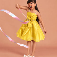 A young girl with dark hair in a purple bow is wearing a bright yellow sleeveless dress with a large bow at the waist, holding a pastel pink and blue ribbon against a solid peach background.