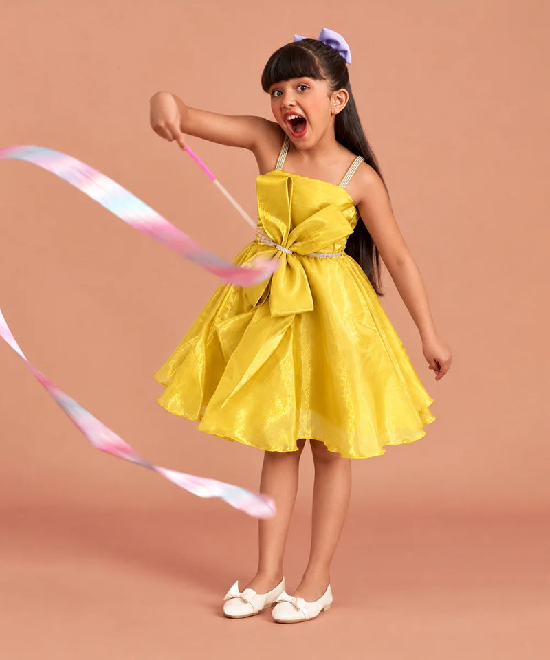A young girl with dark hair in a purple bow is wearing a bright yellow sleeveless dress with a large bow at the waist, holding a pastel pink and blue ribbon against a solid peach background.