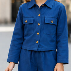A person wearing a cropped blue button-front jacket with flap chest pockets and matching blue pants, photographed outdoors with a blurred background.