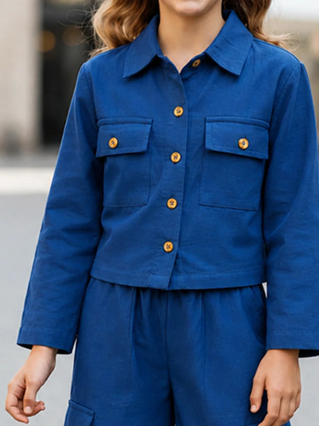 A person wearing a cropped blue button-front jacket with flap chest pockets and matching blue pants, photographed outdoors with a blurred background.