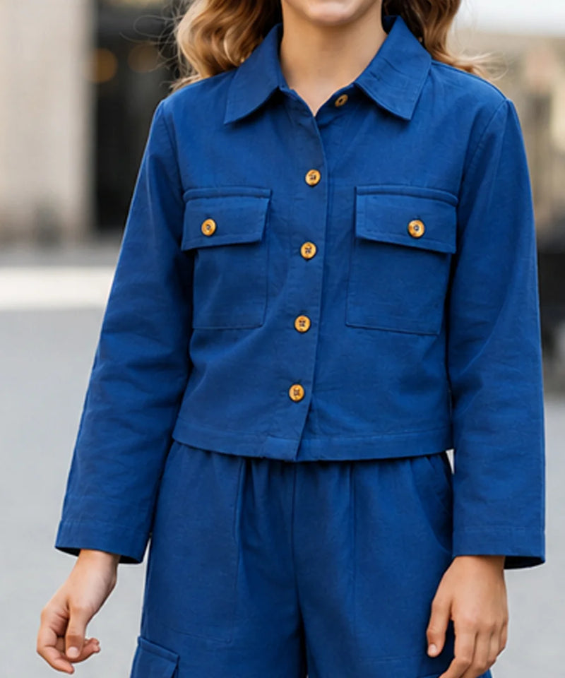 A person wearing a cropped blue button-front jacket with flap chest pockets and matching blue pants, photographed outdoors with a blurred background.