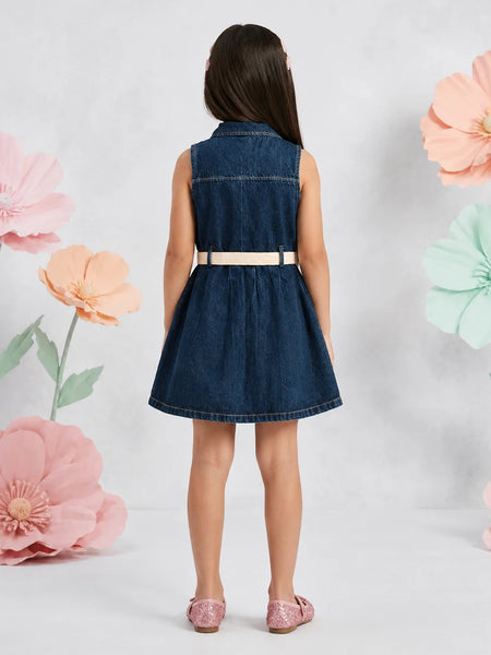 A young girl is shown from the back wearing a sleeveless dark blue denim dress with a beige belt and pink shoes, standing against a white background decorated with large pastel paper flowers.