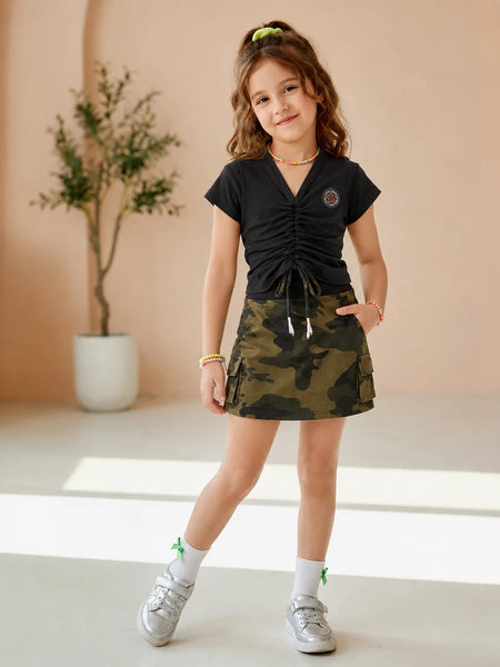 A young girl stands in a studio setting wearing a black ruched short-sleeve top and a green camouflage mini skirt. She is styled with white socks, silver sneakers, and a gold necklace.