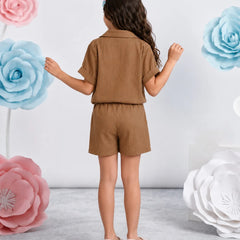 A young girl is shown from behind wearing a matching brown short-sleeve collared shirt and shorts set. She is standing in a studio decorated with large pink, blue, and white paper flowers against a light gray wall.