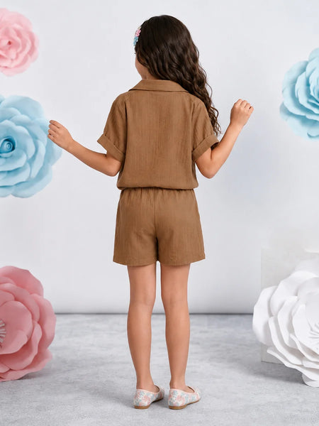 A young girl is shown from behind wearing a matching brown short-sleeve collared shirt and shorts set. She is standing in a studio decorated with large pink, blue, and white paper flowers against a light gray wall.