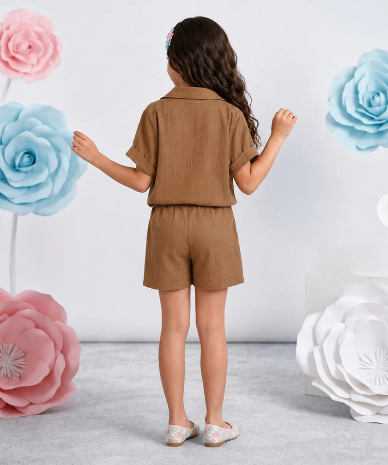 A young girl is shown from behind wearing a matching brown short-sleeve collared shirt and shorts set. She is standing in a studio decorated with large pink, blue, and white paper flowers against a light gray wall.