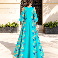 A woman stands with her back to the camera wearing a turquoise blue Anarkali gown decorated with gold and purple floral patterns, set against a stone courtyard background.