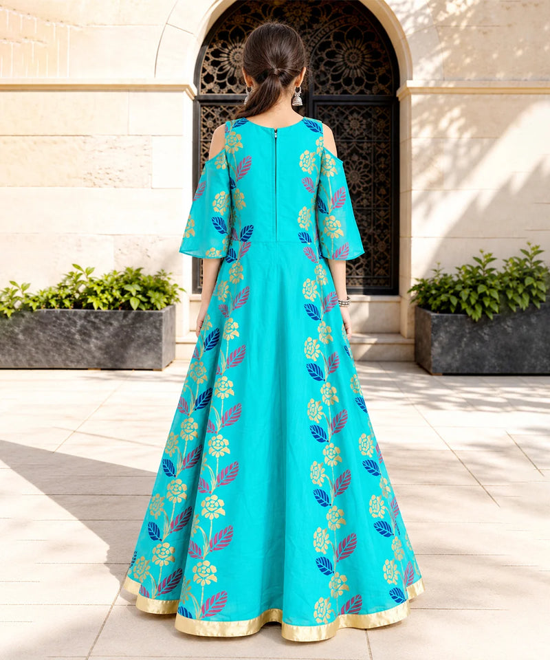 A woman stands with her back to the camera wearing a turquoise blue Anarkali gown decorated with gold and purple floral patterns, set against a stone courtyard background.