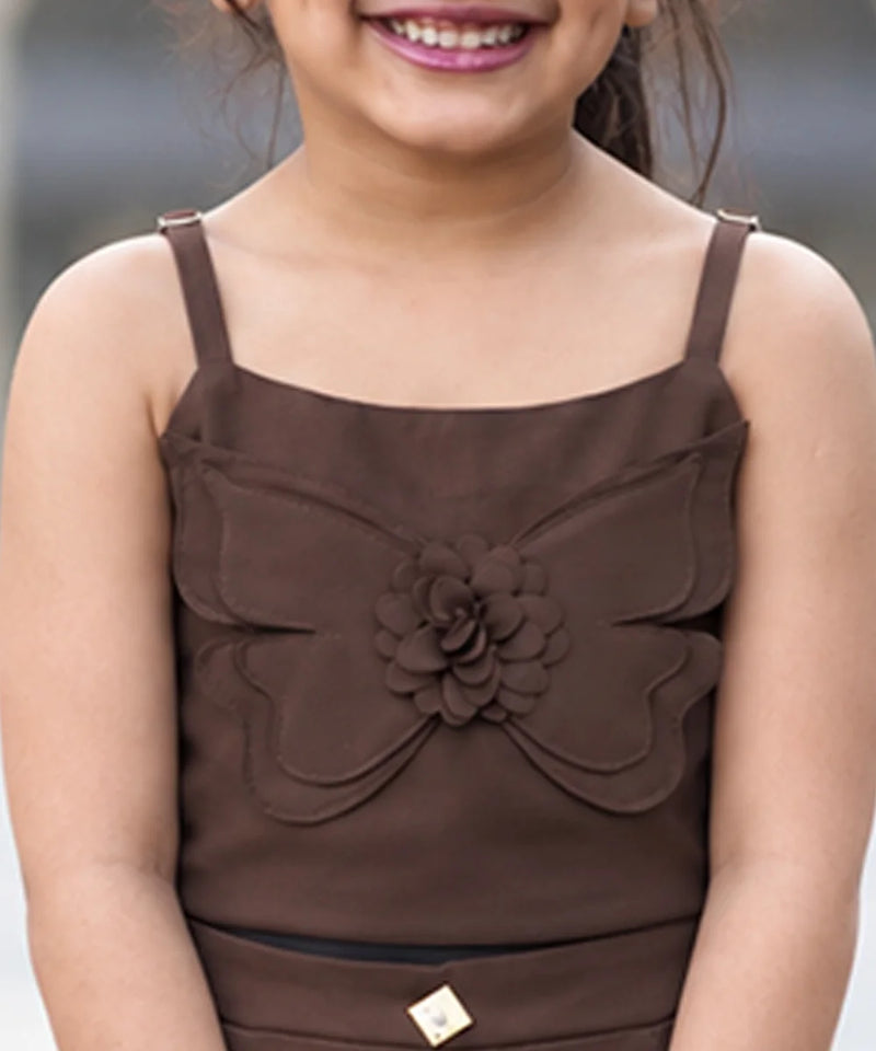 A child model shown from behind wearing a brown two-piece outfit consisting of a smocked camisole top and matching wide-leg trousers, standing in an outdoor plaza.