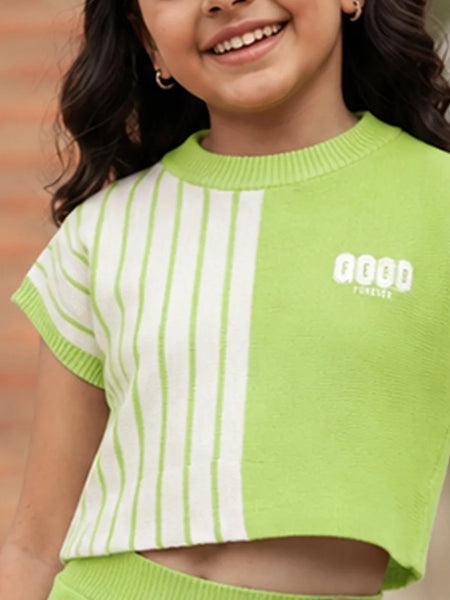 A smiling girl wearing a lime green and white striped short-sleeve knit crop top with a white graphic logo on the chest, set against a blurred brick wall background.