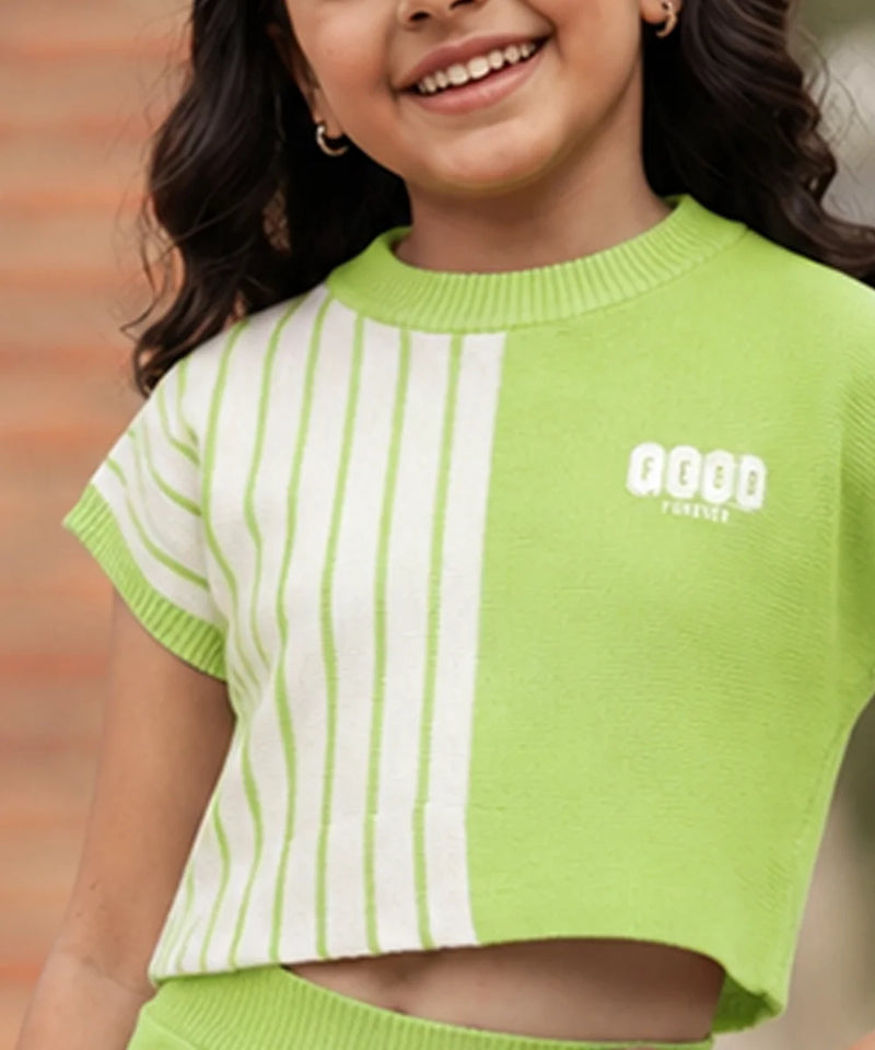 A smiling girl wearing a lime green and white striped short-sleeve knit crop top with a white graphic logo on the chest, set against a blurred brick wall background.
