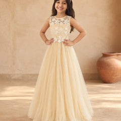 A young girl stands in a warm, rustic indoor setting wearing a formal cream or champagne-colored gown with a lace bodice and a full tulle skirt.
