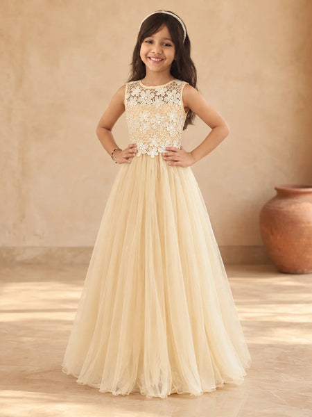 A young girl stands in a warm, rustic indoor setting wearing a formal cream or champagne-colored gown with a lace bodice and a full tulle skirt.