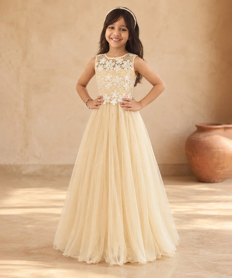 A young girl stands in a warm, rustic indoor setting wearing a formal cream or champagne-colored gown with a lace bodice and a full tulle skirt.