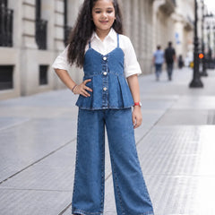 A young girl stands on a city sidewalk wearing a blue denim two-piece outfit consisting of a sleeveless button-front peplum top and matching wide-leg pants, layered over a white short-sleeve collared shirt.