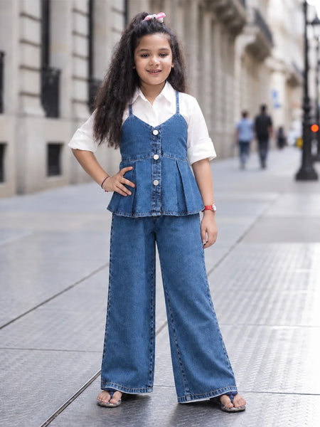 A young girl stands on a city sidewalk wearing a blue denim two-piece outfit consisting of a sleeveless button-front peplum top and matching wide-leg pants, layered over a white short-sleeve collared shirt.