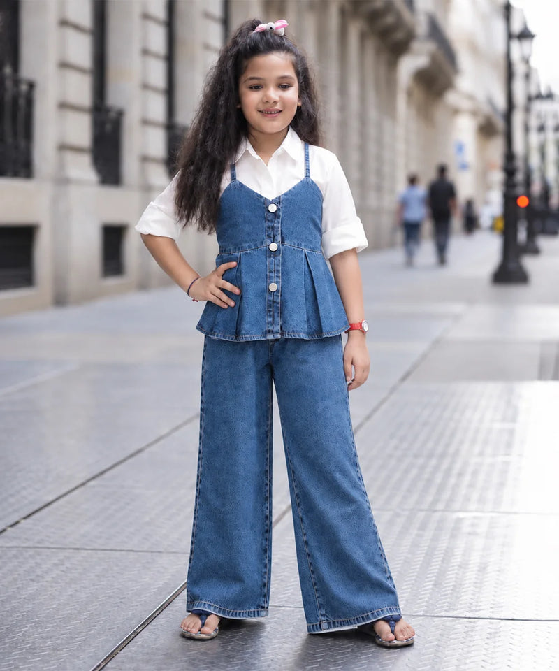 A young girl stands on a city sidewalk wearing a blue denim two-piece outfit consisting of a sleeveless button-front peplum top and matching wide-leg pants, layered over a white short-sleeve collared shirt.
