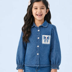 A young girl with long dark hair and pink hair clips is smiling, wearing a blue denim button-up shirt with a decorative bow patch on the chest pocket, paired with blue jeans, against a light blue studio background.