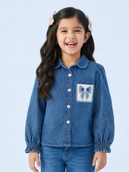 A young girl with long dark hair and pink hair clips is smiling, wearing a blue denim button-up shirt with a decorative bow patch on the chest pocket, paired with blue jeans, against a light blue studio background.